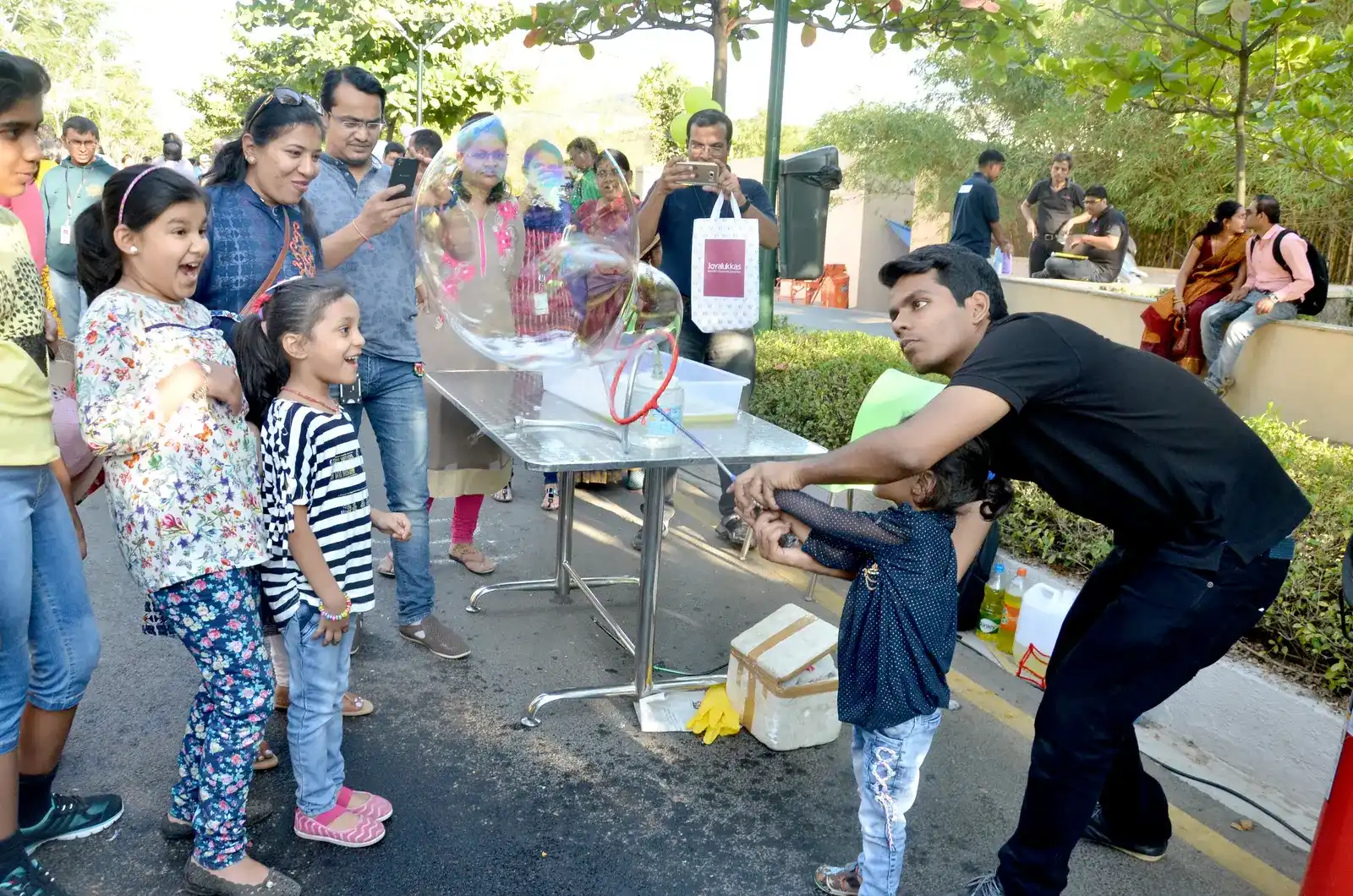 Families gathered on a tree-lined campus road, children delighted as a giant bubble forms at a MakerMasti demonstration